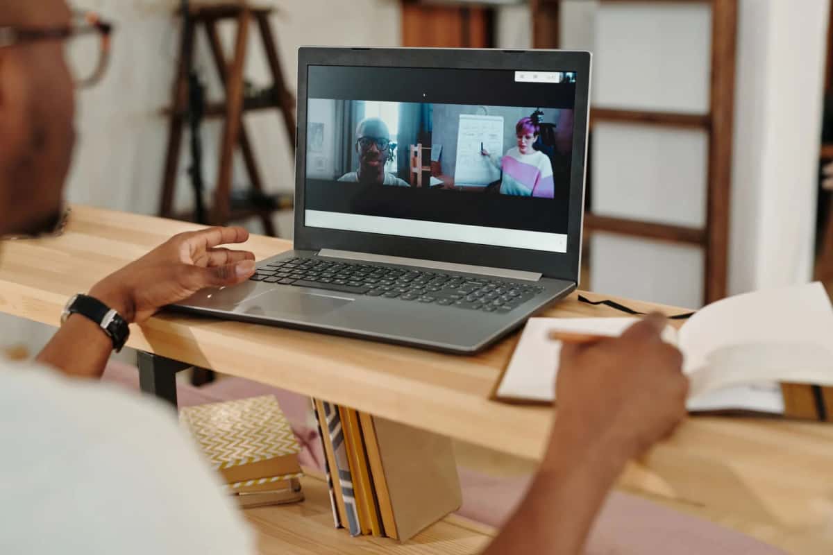 Une personne en visioconférence sur un ordinateur portable, un carnet ouvert sur le bureau en bois