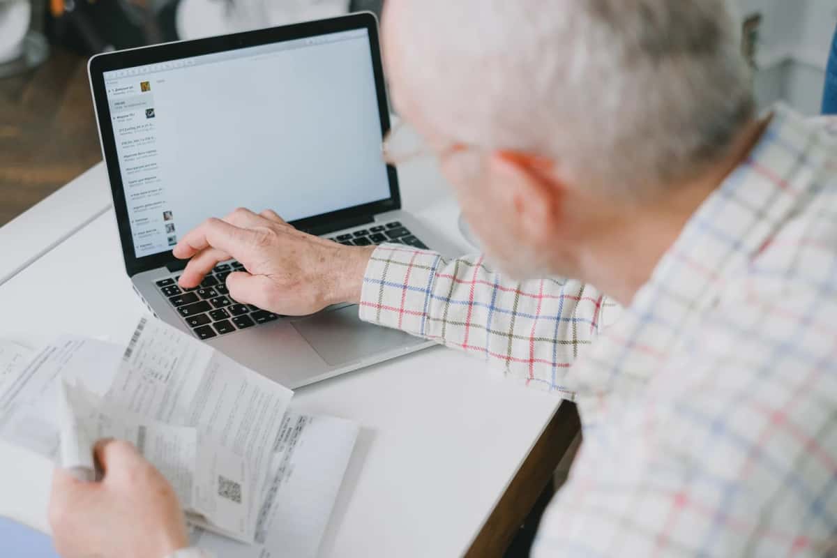 Un retraité consulte des documents papier devant son ordinateur portable pour préparer un dossier en ligne
